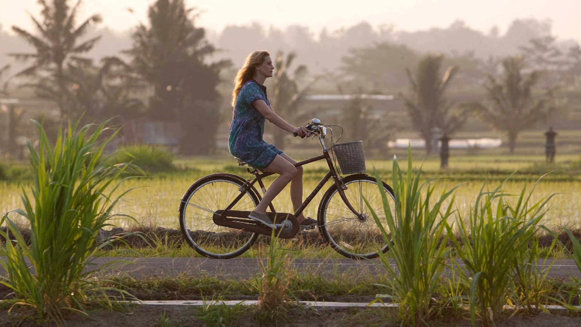 Scene of Elizabeth riding a bicycle through the rural landscape of Bali at sunset, surrounded by rice fields and palm trees.